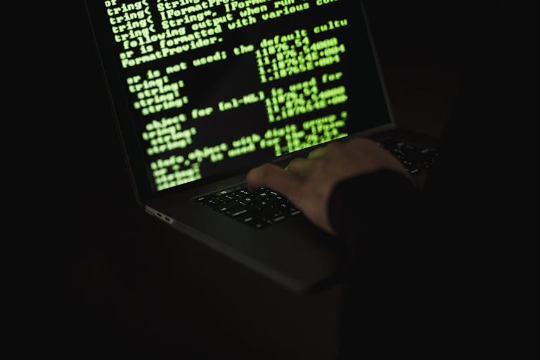 Silhouette of a hacker typing on a laptop in the dark with green code on screen.